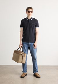 Mornarica polo majica s prugastim ovratnikom, traperice, smeđe cipele i bež torba. Model nosi sunčane naočale i stoji naslonjen na svijetlu pozadinu.