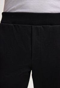 Schwarze Sporthosen mit glatter Textur, ausgestattet mit einem gerippten Bund und dezenten Falten an der Vorderseite für zusätzlichen Komfort und Stil.