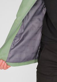 Lichtgroene leren jas met een soepel grijs interieur, voorzien van een ritsluiting en een subtiel merkpatroon op de voering.