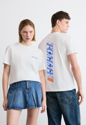 Frau steht frontal, trägt weißes Tommy Jeans T-Shirt und Jeansrock, Mann seitlich gewandt, zeigt blau-orangefarbenes Tommy-Logo auf der Rückseite eines weißen T-Shirts.