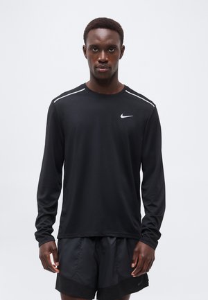 Homme portant un t-shirt Nike noir à manches longues et un short noir, debout devant un fond clair uni, face à l'objectif.