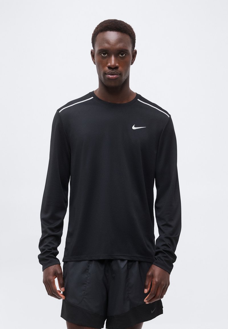 Άνδρας φοράει μαύρο μακρυμάνικο μπλουζάκι Nike και μαύρο σορτς, στέκεται μπροστά σε απλό φωτεινό φόντο, κοιτάζοντας μπροστά.