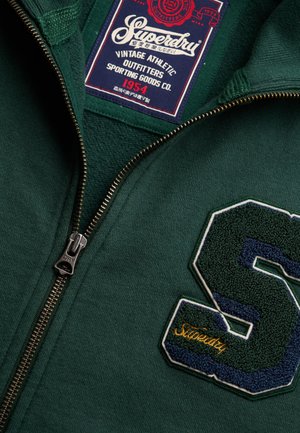 Grøn sweatshirt med lynlås med stort tekstureret navy- og grønt "S"-mærke og synligt Superdry-mærke inde i kraven.