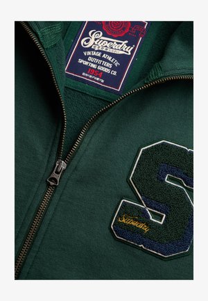Zielona bluza zapinana na zamek z dużą, fakturowaną naszywką "S" w kolorze granatowo-zielonym oraz widoczną metką marki Superdry wewnątrz kołnierza.