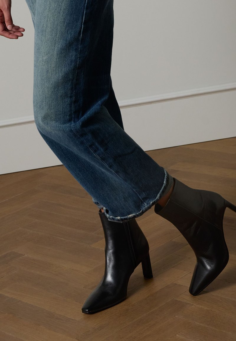 Schwarze Leder-Stiefeletten mit spitzer Schuhspitze und schlankem, hohem Absatz, kombiniert mit blauen Jeans mit ausgefranstem Saum.