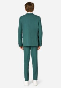 Completo teal con una vestibilità slim, caratterizzato da un risvolto a punta e pantaloni su misura. Il materiale sembra liscio, abbinato a sneakers bianche.