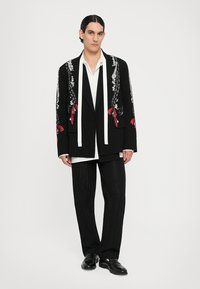 Blazer nero con intricati ricami bianchi e rossi, dotato di lunghe cravatte bianche, abbinato a una camicia bianca e pantaloni neri strutturati.
