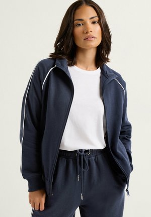 Joven mujer que lleva una chaqueta deportiva azul marino con cremallera y ribetes blancos sobre una camiseta blanca y pantalones azul marino a juego con cordón, manos en los bolsillos.