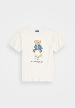 T-shirt blanc à manches courtes avec un dessin d'un ours portant une veste bleue, un pantalon blanc et tenant un sac filet. Texte en dessous : « Polo Bear ».