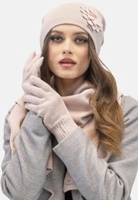 Un bonnet en tricot rose avec un détail de fleur en tissu, des gants assortis et une écharpe légère, associé à un manteau gris avec une texture douce.