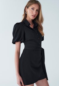 Vestido camisa negro con mangas abullonadas, cuello con solapa, botones al frente y detalle de cinturón ajustable, con una textura de tela suave.