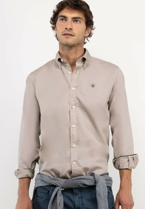 Hombre con camisa beige de botones con las mangas remangadas, un suéter gris atado alrededor de la cintura y pantalones azules, mirando ligeramente hacia arriba.