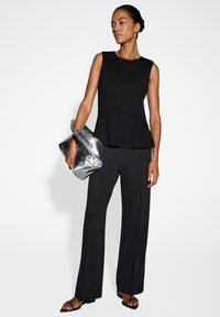 Zwart mouwloos peplumtop gecombineerd met wijdbesneden zwarte broek. Het model houdt een gekreukeld zilveren clutch-tas vast. Glad materiaal, minimalistisch ontwerp.