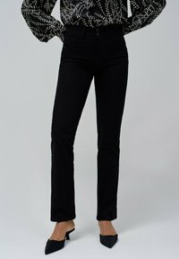 Jeans negros de corte slim con una cintura de altura media, que cuentan con cinco bolsillos y detalles de costura sutiles. Combinados con mules negros puntiagudos.