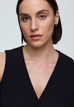 Srebrny naszyjnik z okrągłym wisiorkiem noszony na czarnym bezrękawniku. Wisiorek ma mały przezroczysty kamień na środku.