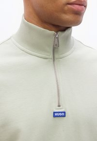 Άνδρας που φοράει ανοιχτό πράσινο ζακέτα με φερμουάρ και ψηλό γιακά, με μπλε ετικέτα λογότυπου "HUGO" κάτω από το φερμουάρ.
