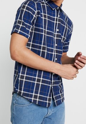 Mand iført kortærmet blåternet knap-op skjorte og lyseblå jeans, justerer ærmerne med hænderne foran hvid baggrund.