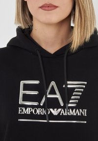 Felpa nera con cappuccio, caratterizzata da un logo argento con il testo "EA7" e "EMPORIO ARMANI". Il tessuto sembra morbido, con un cappuccio regolabile e una vestibilità comoda.