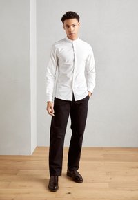 Camisa blanca de manga larga con cuello de banda, combinada con pantalones negros y zapatos marrones de tipo mocasín, sobre un fondo claro.