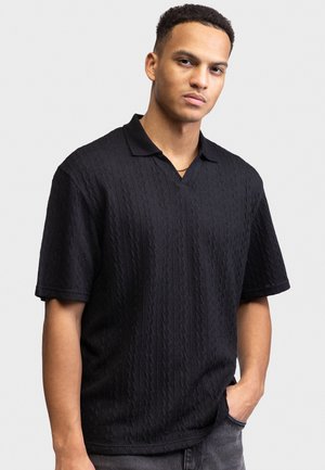 Νεαρός άνδρας με κοντά σγουρά μαλλιά, φορώντας μαύρο πόλο με υφή και κοντά μανίκια και γκρι τζιν, όρθιος με το χέρι στην τσέπη.