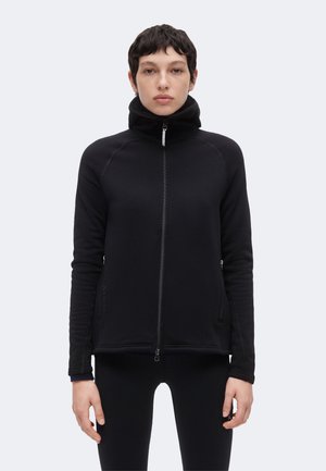 Femme aux cheveux courts et foncés portant une veste noire zippée à col montant et un pantalon noir, debout devant un fond clair uni.