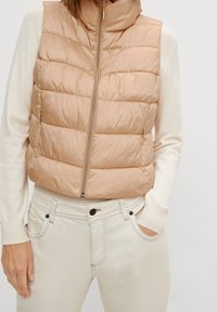 Béžová prošívaná vesta s horizontálním prošitím, vysokým límcem a zapínáním na zip. Nositelná přes krémový svetr, doplněná světlými džínami.