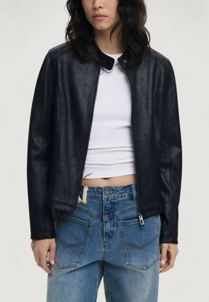 Sort læderjakke med fitted design, lynlås og høj krave. Båret over en hvid skjorte og parret med slidte blå jeans.
