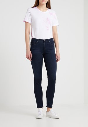 Vrouw draagt donkerblauwe skinny jeans, witte sneakers en een wit T-shirt met lichtroze tie-dye accenten, staand tegen een effen achtergrond.