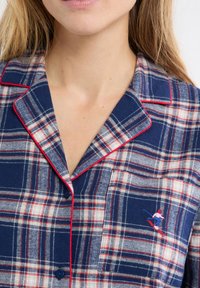 Camicetta per pigiama a quadri blu navy con accenti bianchi e rossi; presenta una patta con bottoni e una figura da sci ricamata sulla tasca. Materiale in cotone.