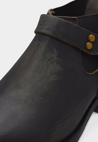 Μαύρο δερμάτινο μποτάκι αστραγάλου με διακοσμητικό λουράκι και δύο μεταλλικά τρουκς σε χρυσό τόνο κοντά στο άνοιγμα του αστραγάλου.