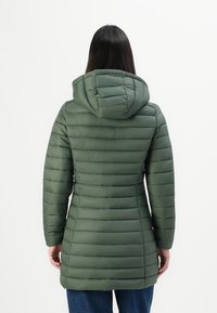 Vrouw met lang donker haar, gekleed in een getailleerde groene gewatteerde winterjas met capuchon en blauwe jeans, gezien vanaf de achterkant op een witte achtergrond.