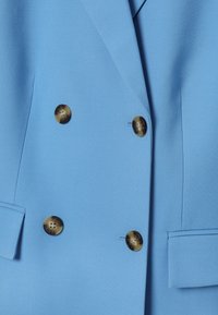 Blazer bleu clair à double boutonnage avec des boutons en écaille de tortue marron et des poches à rabat de chaque côté.