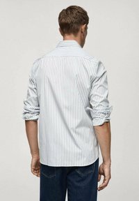 Camicia a righe bianca e blu, con maniche arrotolate, tessuto leggero, colletto classico e orlo curvo. Indossata con jeans scuri.