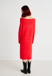 Abito rosso a maglia off-shoulder con maniche lunghe, di lunghezza al ginocchio, con scollo a coste. Indossato con stivali neri a punta alla caviglia.