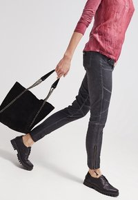 Zwarte denimspijkerbroek met zijritsen, gedragen met een gestructureerde rode lange mouwen top; gecombineerd met een zwarte suède tas en zwarte veterschoenen.