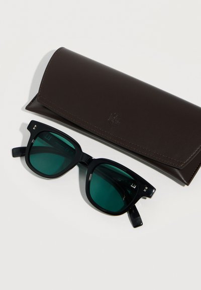 Γυαλιά ηλίου με μαύρο πλαίσιο και φακούς με πράσινη απόχρωση δίπλα σε θήκη γυαλιών από σκούρο καφέ δέρμα πάνω σε ανοιχτόχρωμη επιφάνεια.