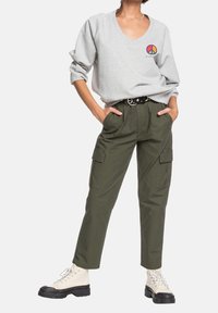 Sudadera gris de cuello en V con un colorido símbolo de paz; pantalones cargo ajustados en color oliva con bolsillos; botas beige con cordones y suelas negras.