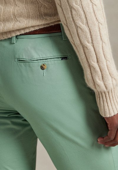 Šviesiai žalios kelnės iš lygaus audinio, su užsegamu galiniu kišene ir mažu etiketės detalizavimu. Dėvimos kartu su kremine pynimo megztine.