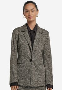 Kvinna med mörkt hår i en knut som bär guldörhängen, en kavaj med fiskbensmönster och en svart skjorta mot en vit bakgrund.