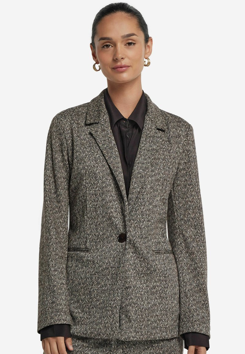 Kvinna med mörkt hår i en knut som bär guldörhängen, en kavaj med fiskbensmönster och en svart skjorta mot en vit bakgrund.