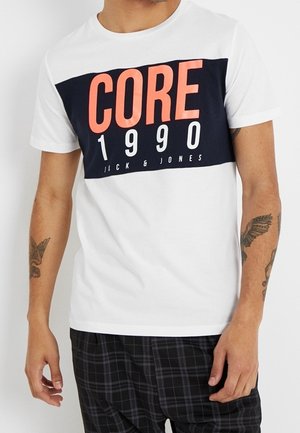 Homme portant un t-shirt blanc avec un panneau rectangulaire noir et un texte orange "CORE 1990 Jack & Jones", associé à un pantalon à carreaux noir et gris.