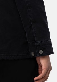 Veste noire en tissu texturé, mettant en avant un gros plan de la manche montrant deux boutons-pression en métal et des détails de couture.