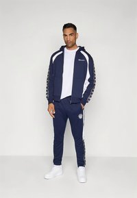 Fato de treino azul-marinho com detalhes brancos, composto por um casaco com capuz e fecho de correr e calças com cintura elástica. Material parece macio; inclui detalhes do logótipo.