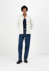 Lichtbeige overhemd met knopen, marineblauw tanktop, donkerblauwe high-waisted jeans en zwarte schoenen. Glad materiaal, oversized pasvorm en ronde knopen.