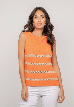 Femme aux longs cheveux noirs portant un haut orange sans manches avec des rayures horizontales beige et un pantalon blanc, debout devant un fond uni.