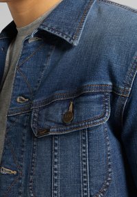 Jeansjacka i mörkblått, med en spetsig krage, bröstficka med en metallknapp och detaljer i kontrasterande sömmar.