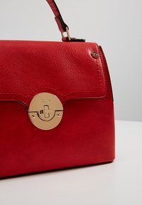 Sac à main en cuir rouge avec une fermeture circulaire dorée et une sangle courte, reposant sur une surface blanche sur un fond clair uni.