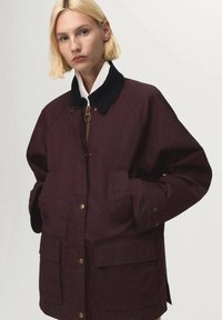 Chaqueta burdeos con cuello de pana, cremallera dorada y bolsillos frontales. Ajuste casual, textura suave y detalles de botones en los puños.