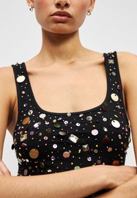 Frau trägt ein schwarzes, geripptes Tanktop, verziert mit mehrfarbigen Pailletten und Perlen, zeigt Dekolleté, Schultern und verschränkte Arme.