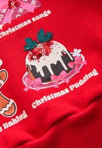 Tessuto rosso con una stampa di budino di Natale con glassa bianca, agrifoglio verde e paillettes rosse, accompagnato da fiocchi rosa e testi.